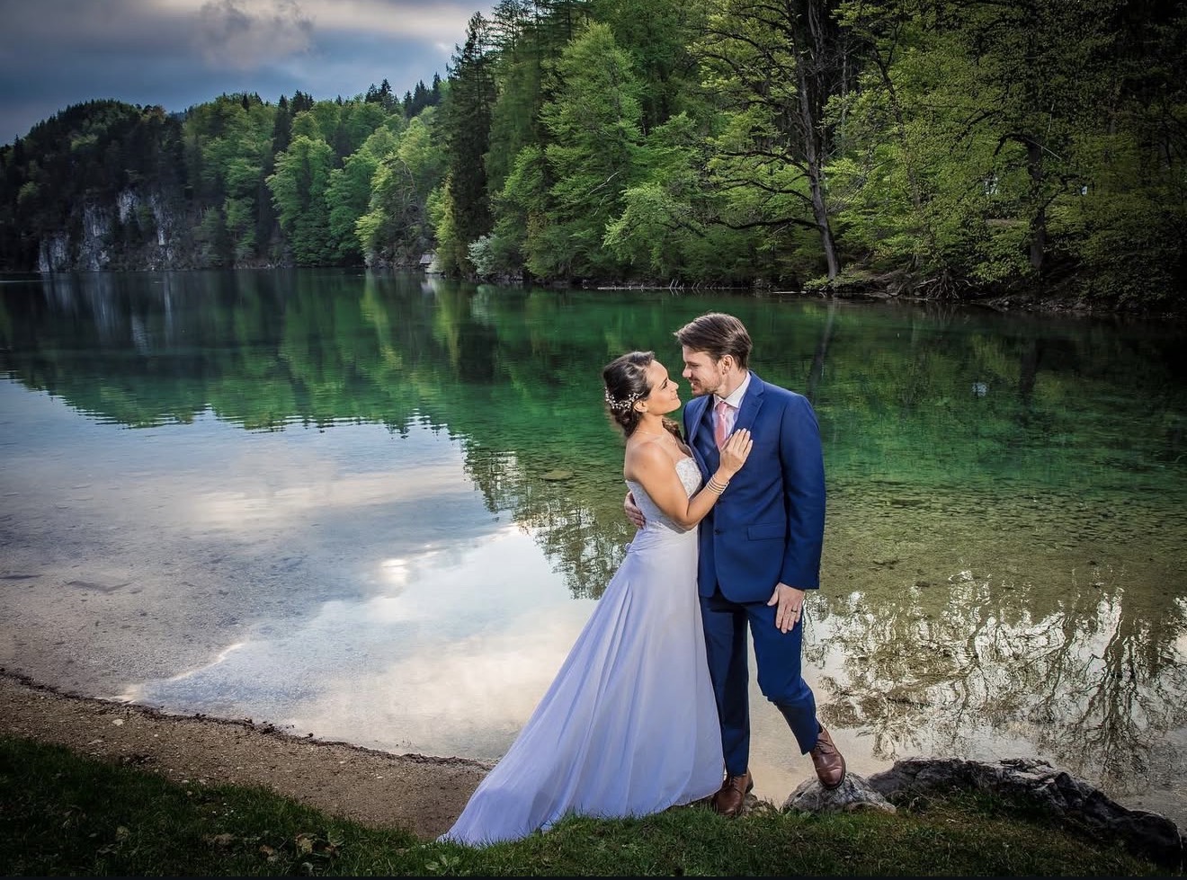 Novios abrazados a orillas de un lago entre bosques y montañas