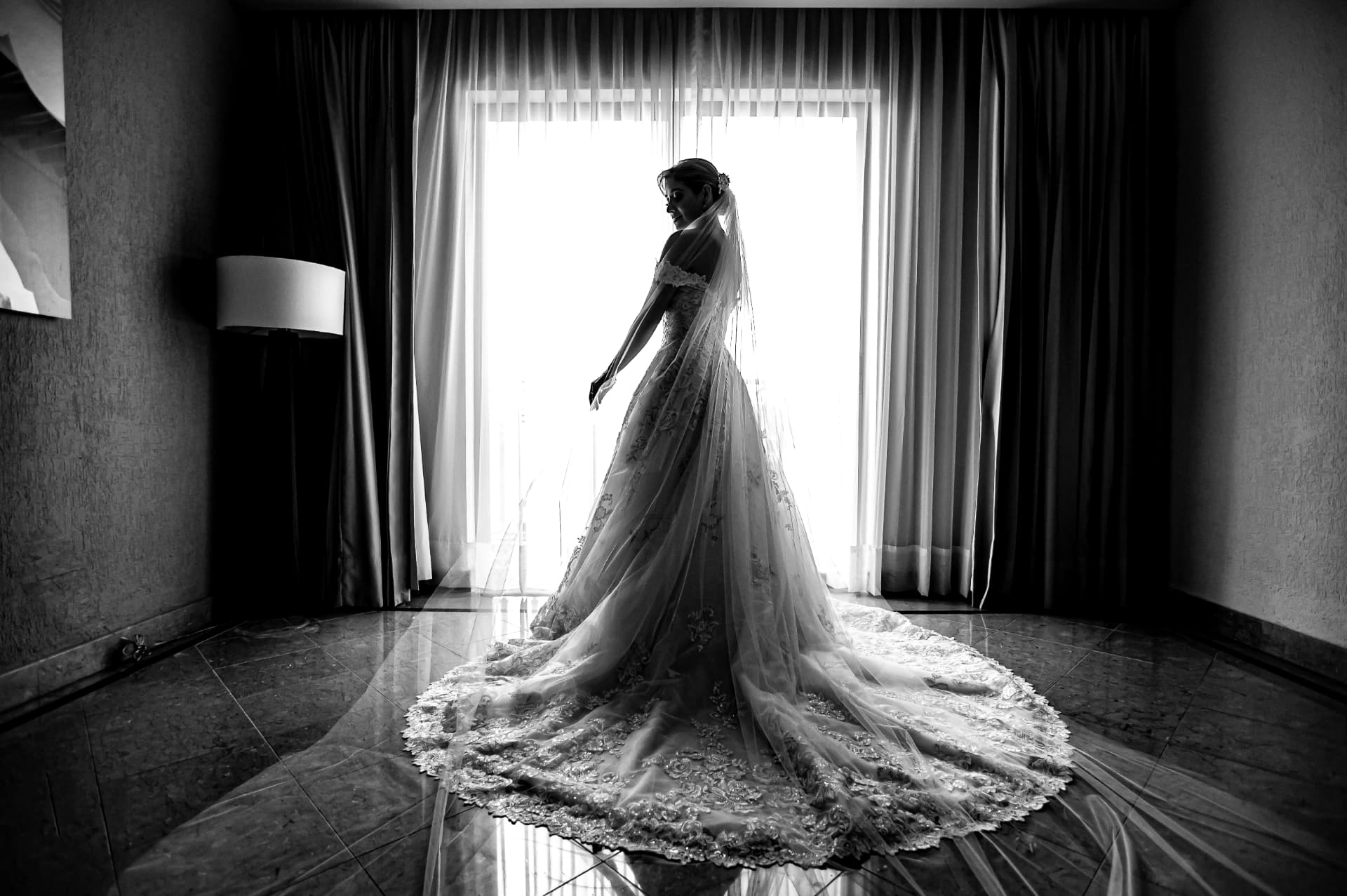 Silueta de novia junto a una ventana mostrando la cauda de su vestido en blanco y negro