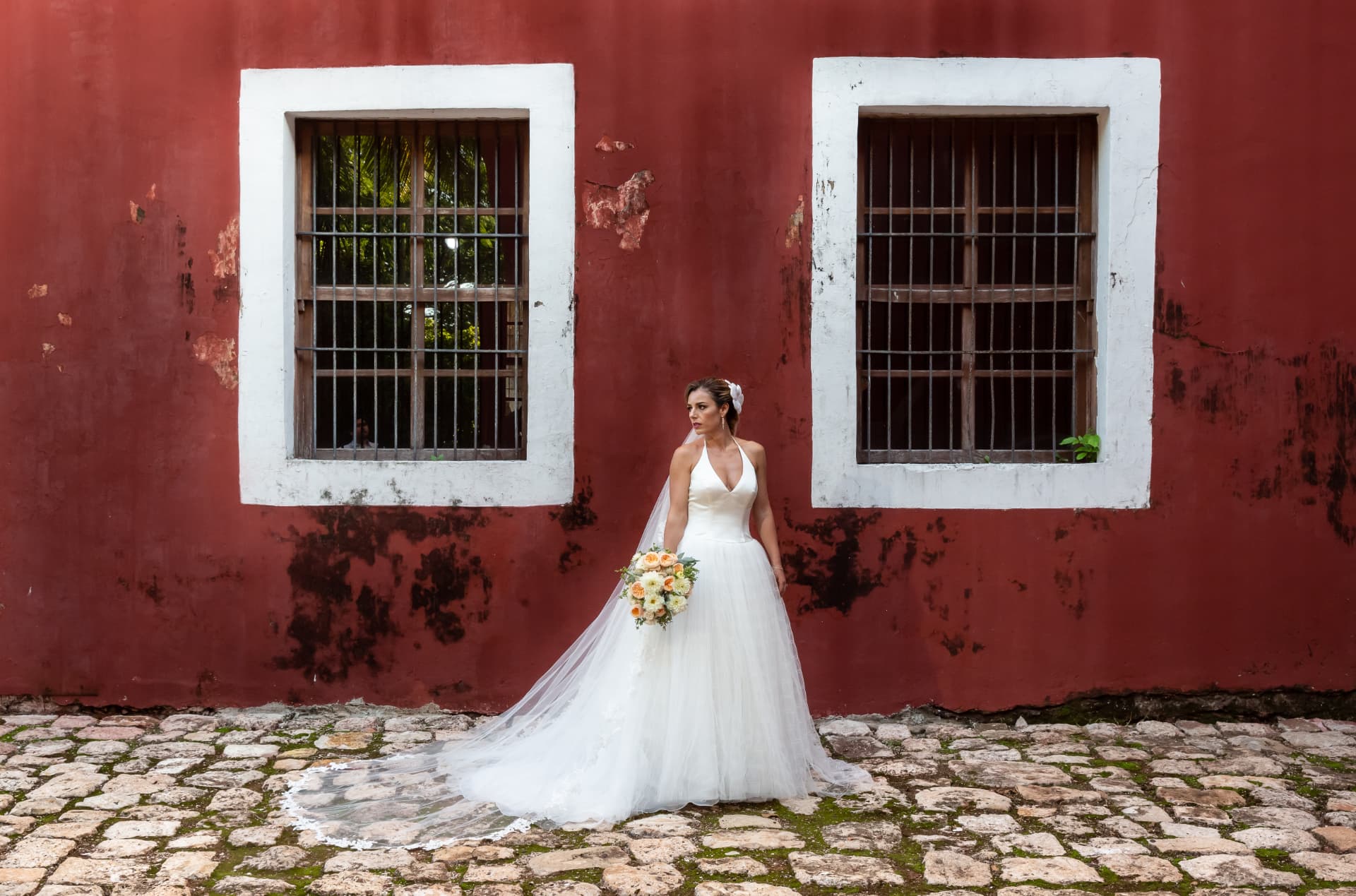 Novia con velo largo frente a un muro colonial rojo con ventanas blancas