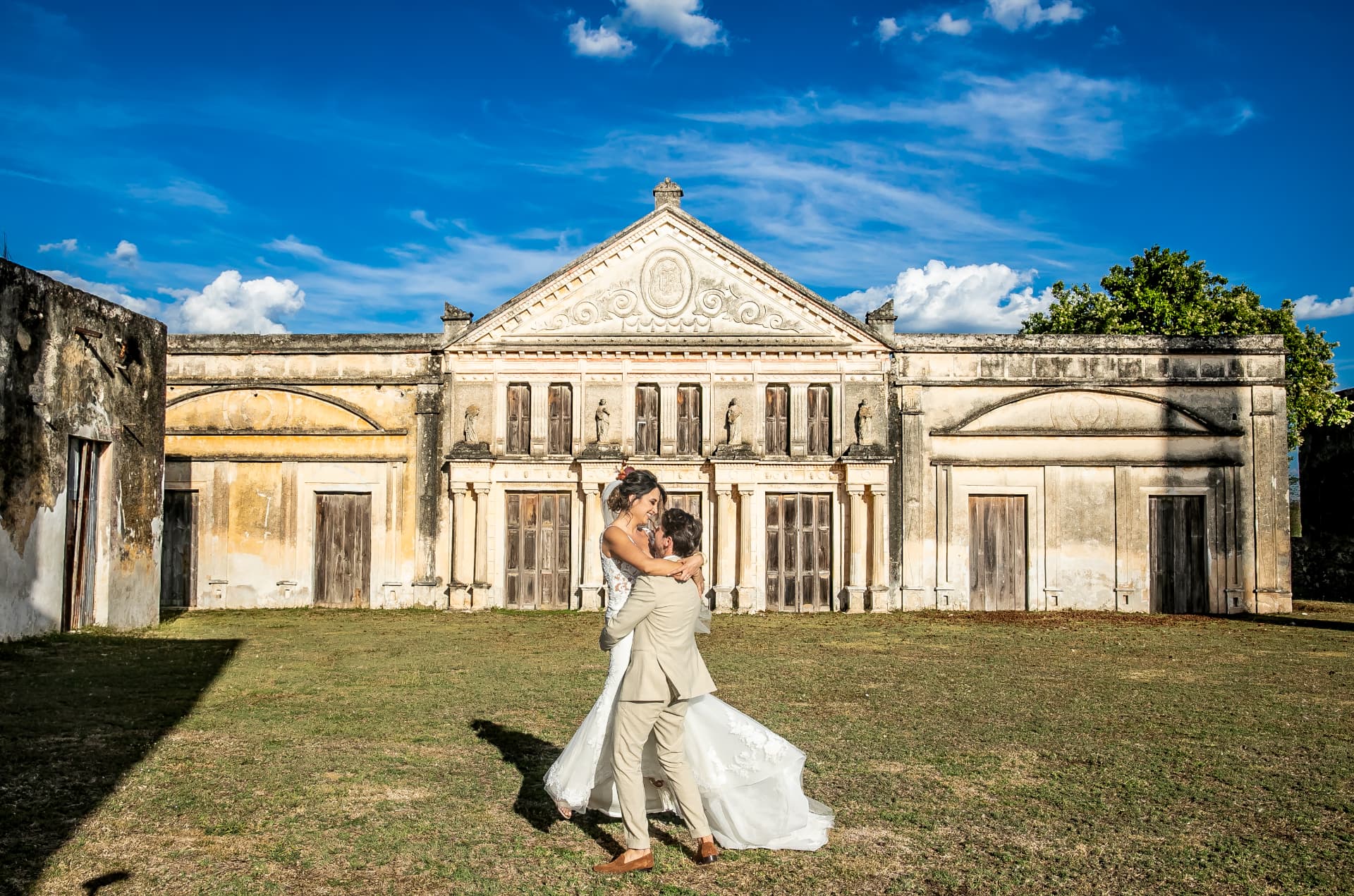 Novios abrazados frente a la fachada histórica de una hacienda bajo un cielo azul