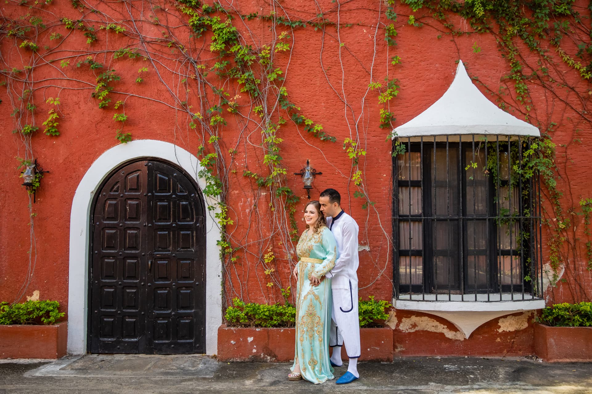 Pareja con atuendo tradicional frente a un muro colonial rojo con enredaderas