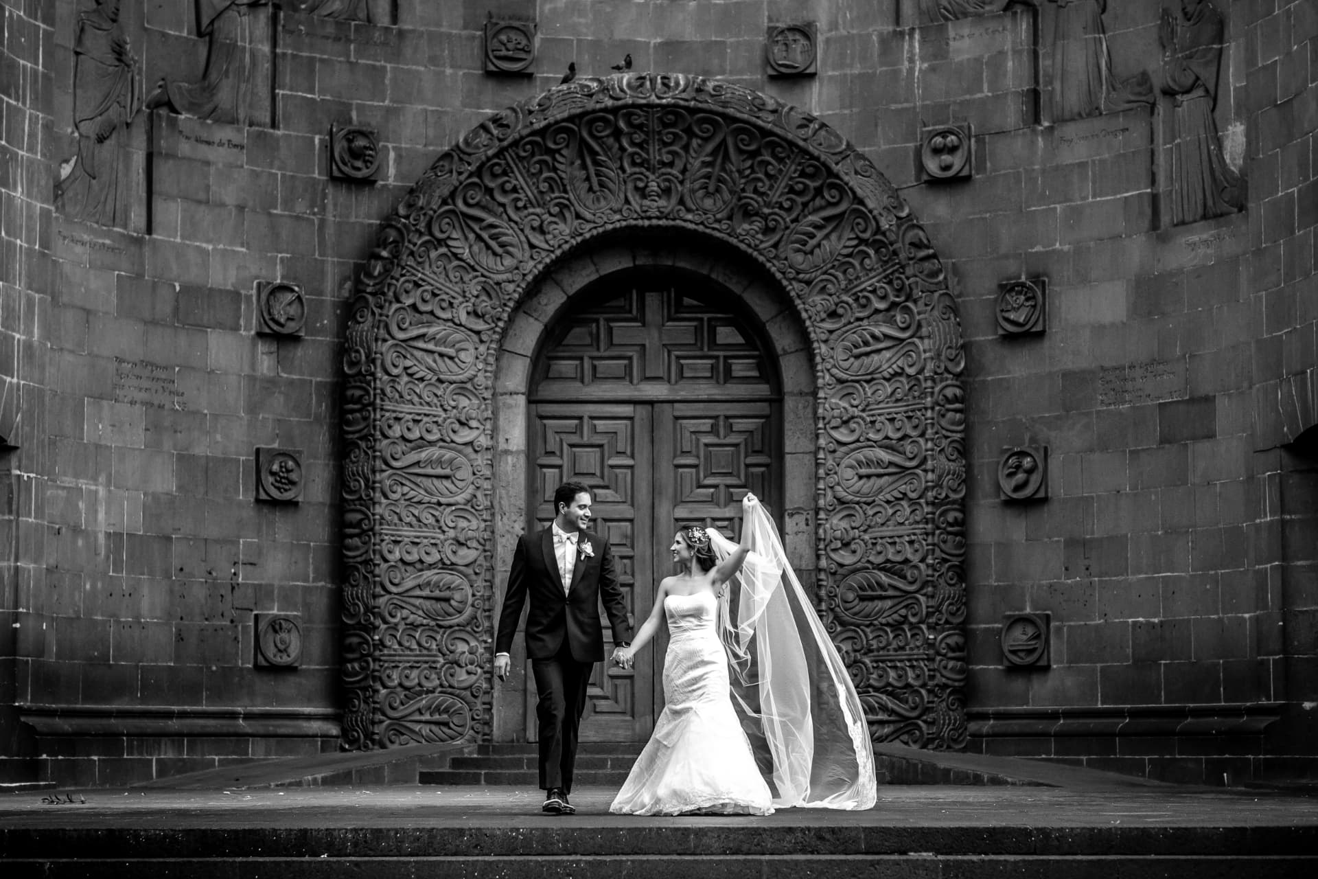 Novios caminando frente a una portada histórica tallada, fotografía de boda en blanco y negro